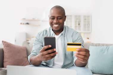 Çevrimiçi alışveriş. Kredi kartı ve akıllı telefonlu neşeli Afro-Amerikalı adam evdeki koltukta oturuyor.