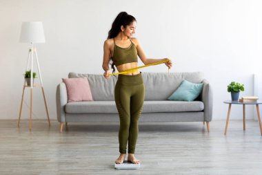 Güzel bir Hintli kadın, terazinin üzerinde dururken belini ölçüyor, evde mükemmel vücut parametrelerine sahip.