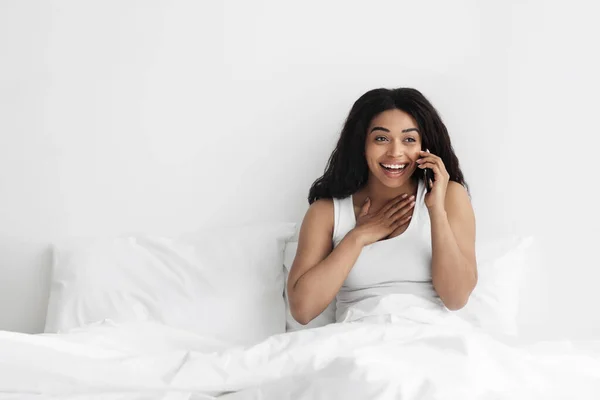 Buenas noticias matutinas. Mujer afroamericana feliz hablando en el teléfono inteligente mientras está sentado en la cama en el dormitorio de luz, espacio vacío — Foto de Stock