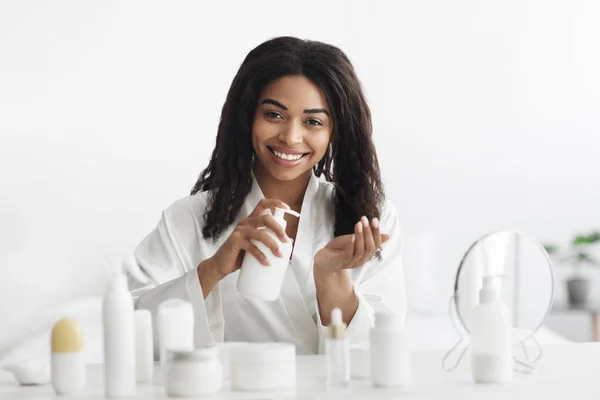 Glimlachende zwarte vrouw met behulp van reinigingsmiddel of lotion, reiniging van haar gezicht of hydraterende huid, zitten in de slaapkamer — Stockfoto