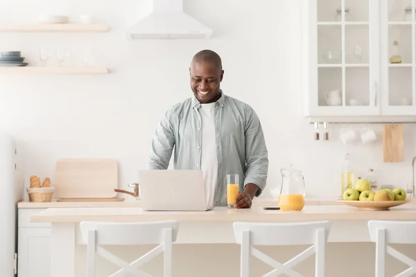 Positive black mature man reading online news on laptop and drinking orange juice in kitchen, enjoying morning at home — Stock Photo, Image
