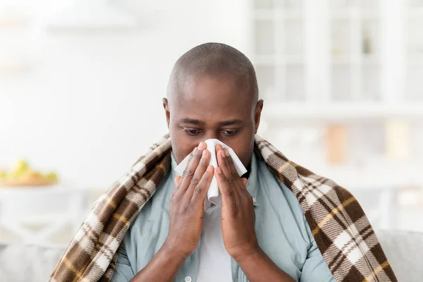 Hombre afroamericano maduro enfermo cubierto de manta que sufre de frío o alergias, sonándose la nariz, sentado en casa — Foto de Stock