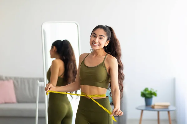 Sorrindo jovem mulher indiana de pé perto do espelho e medindo sua cintura em casa. Perda de peso e dieta — Fotografia de Stock