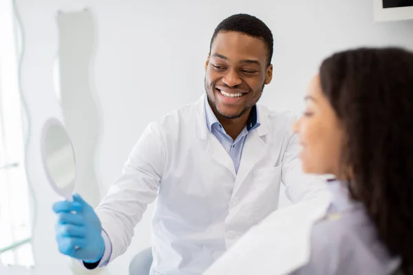 Μαύρος οδοντίατρος γιατρός κρατώντας καθρέφτη, που δείχνει το αποτέλεσμα της θεραπείας των δοντιών στον ασθενή — Φωτογραφία Αρχείου