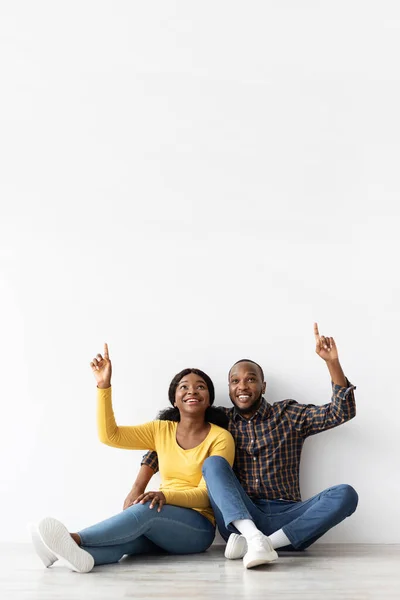 Szczęśliwa afrykańska para siedzi na podłodze przy białym murze i wskazuje — Zdjęcie stockowe
