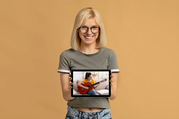 Joven mujer feliz mostrando PC tableta con lección de guitarra en línea en la pantalla sobre fondo de estudio beige —  Fotos de Stock