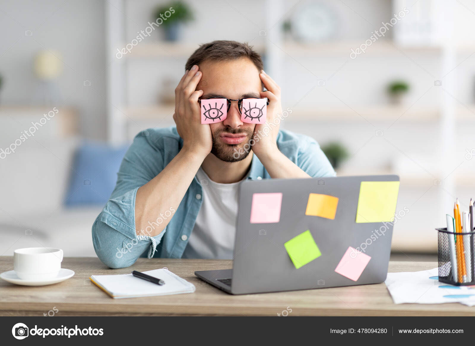Young business man with fake eyes painted on paper stickers yawning at  workplace in office Stock Photo