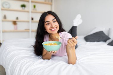 Kilo vermek için dengeli bir diyet. Evde yatakta uzanmış, mısır gevreğini sütle yiyen güzel Hintli bir kadın.