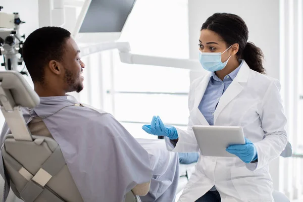 Dentista preto senhora com tablet digital em mãos consultando paciente masculino na clínica — Fotografia de Stock