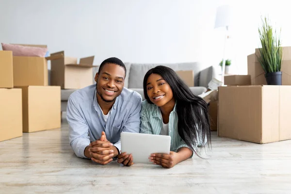Happy black family lying on floor in new apartment with digital tablet among cardboard boxes with belongings — Stock Photo, Image
