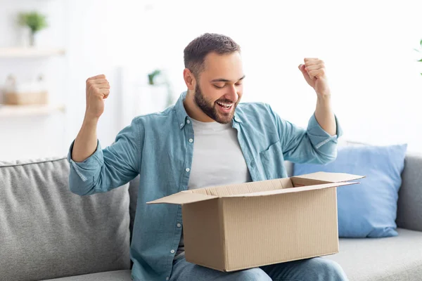 Задоволений чоловічий клієнт відкриває коробку, схвильований службою доставки, отримує замовлення інтернет-магазину, жестикулює YES вдома — стокове фото