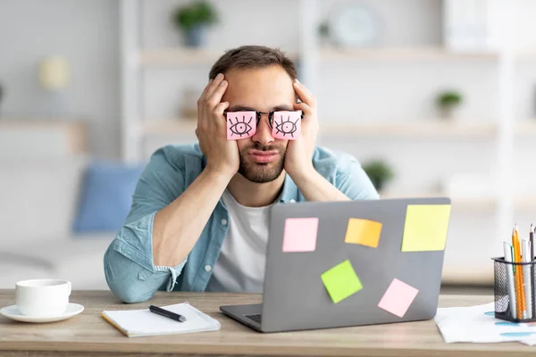 Trött ung man med klistermärken med öppna ögon rita på sina glasögon, sova framför laptop på hemmakontoret — Stockfoto