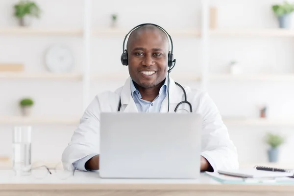 Blog and answer patient questions. Happy african american doctor with headphones and laptop computer at workplace — стоковое фото