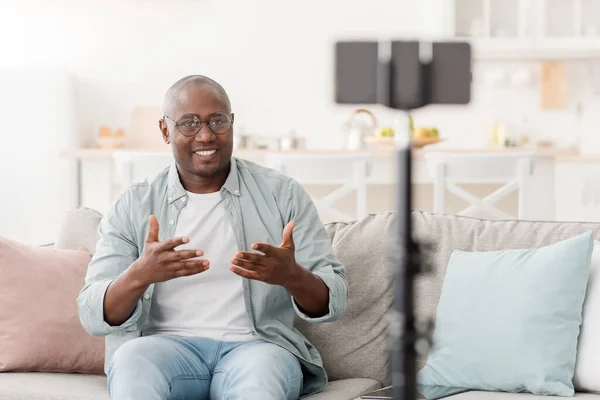 Neşeli, olgun Afrikalı Amerikalı adam evden yayın yapıyor, akıllı telefon ve tripodu kullanıyor, evdeki kanepede oturuyor. — Stok fotoğraf