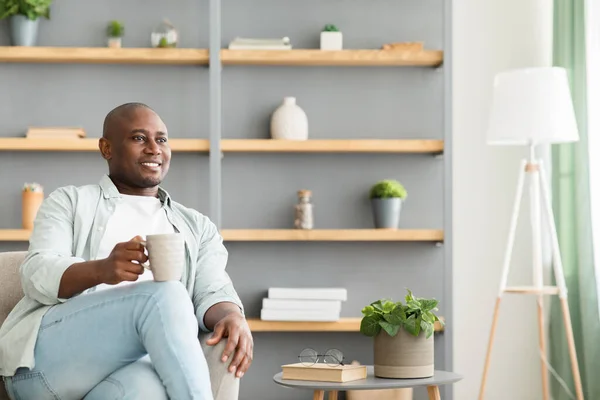 Gülümseyen ve düşünceli Afrikalı Amerikalı adam kahve içiyor, oturma odasındaki koltukta oturuyor, boş yer var. — Stok fotoğraf