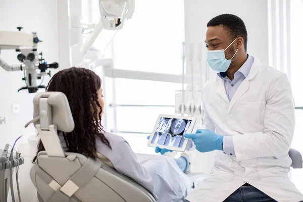 Αρσενικός Μαύρος Οδοντίατρος γιατρός που επιδεικνύει γυναικεία δόντια ασθενών ακτίνων Χ σε ψηφιακό δισκίο — Φωτογραφία Αρχείου