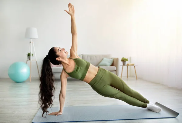 Fit mulher indiana fazendo prancha lateral no tapete esportivo dentro de casa, comprimento total. Conceito de treino de força doméstica — Fotografia de Stock