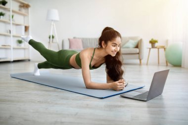 Dirsek tahtasında duran güçlü Hintli kadın, bacağını spor minderinde kaldırıyor, dizüstü bilgisayardaki video dersini takip ediyor.