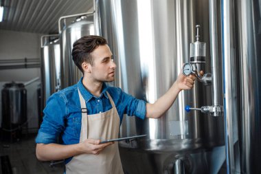 Bira fabrikası işçisi çelik fıçıdaki fermantasyon sürecini kontrol ediyor ve fabrikadaki not defterine bakıyor.