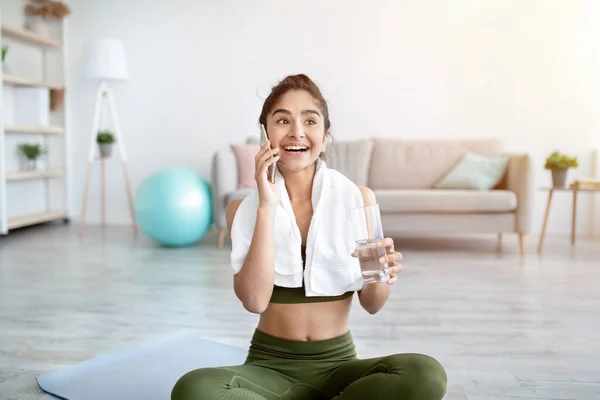 Giovane donna indiana sportiva con un bicchiere d'acqua che parla sul cellulare, dopo aver interrotto l'allenamento a casa — Foto Stock