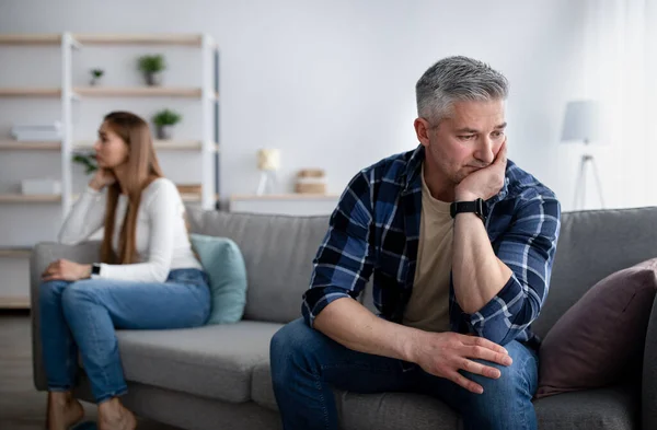 Offended mature man and woman sitting on different sides of sofa after after quarrel indoors, empty space — Stockfoto
