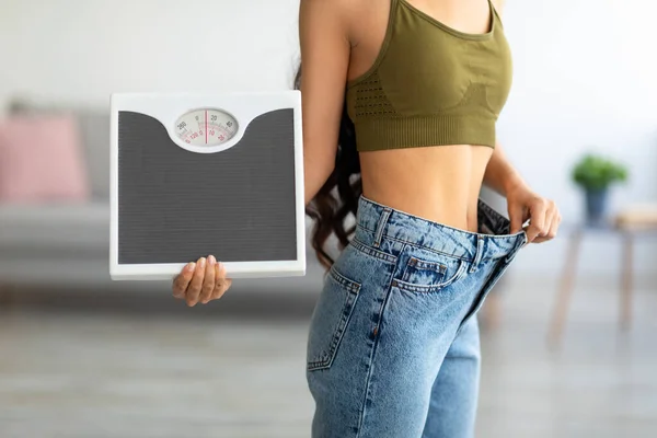 Cropped view of Indian woman in oversized jeans holding scales, impressed with results of weight loss diet — 스톡 사진