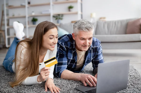 Casais maduros alegres encomendar bens on-line com laptop pc e cartão de crédito, deitado no chão em casa — Fotografia de Stock