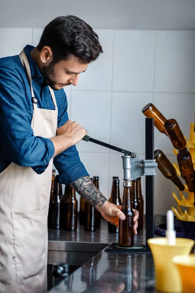 Jovem trabalha em pequena fábrica de cerveja artesanal e tampas garrafa de cerveja — Fotografia de Stock