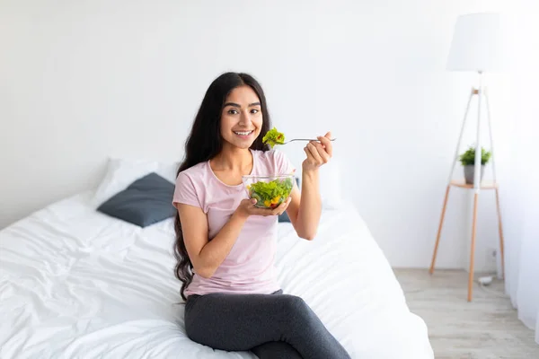 Hälsosam kost koncept. Vacker indisk kvinna äter smaskig grönsakssallad när hon sitter på sängen hemma — Stockfoto