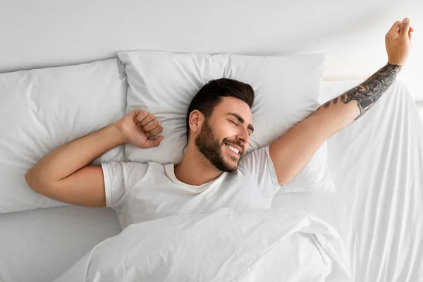 Awakening concept. Happy rested wellslept man waking up in morning and stretching hands, lying in bed, top view — Foto Stock