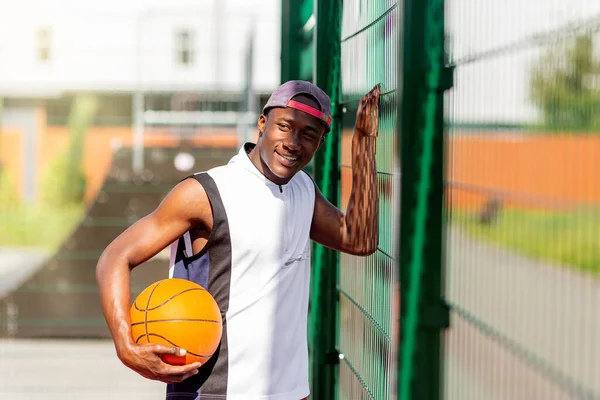 Portrait of millennial African American sportsman with basketball standing near fence at outdoor stadium, copy space — Stok Foto