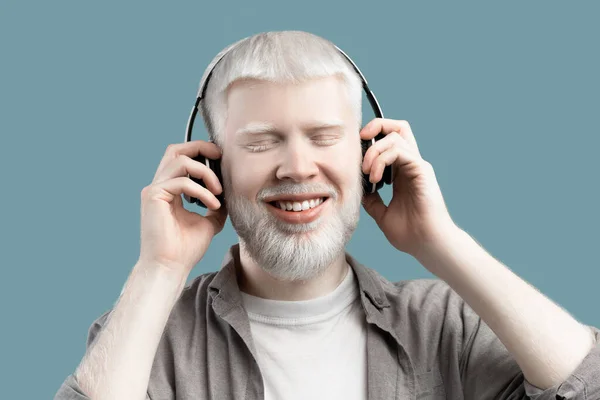 Happy albino man listening music in wireless headphones and touching them on turquoise studio background — Foto de Stock