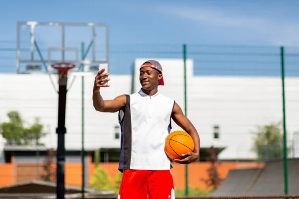 Portrait of black basketball player holding ball, taking selfie on smartphone at outdoor sports arena — Stok Foto