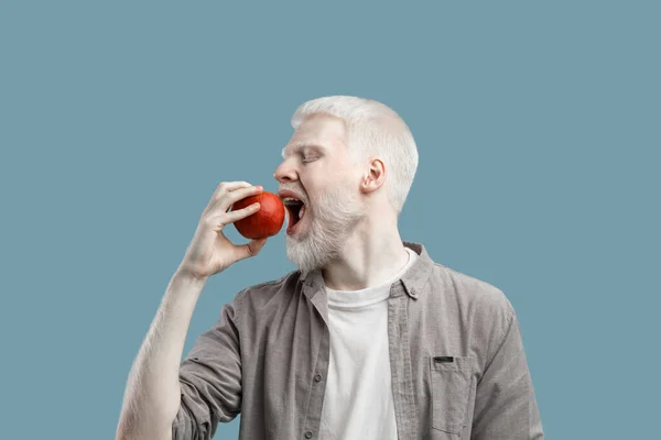 Eat fruits. Young albino man biting fresh red apple, standing over turquoise studio background — Stok Foto