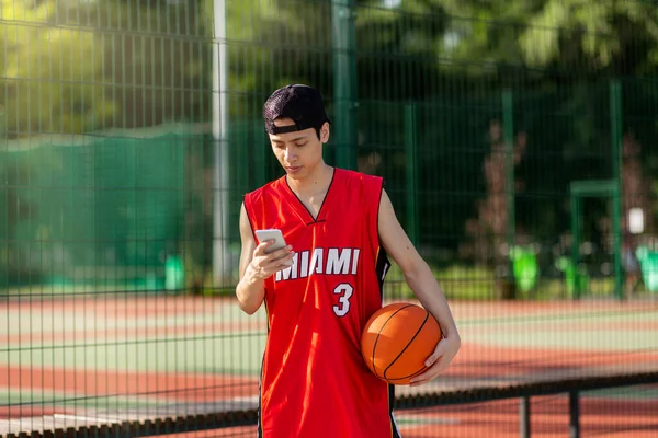 Attractive Asian sportsman checking his smartphone on break from basketball game at outdoor court, free space — Stok Foto