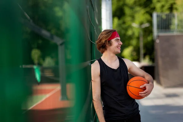 Millennial Caucasian basketball player with ball standing near fence at stadium, empty space — Stok Foto