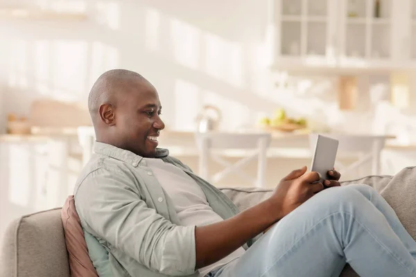 Happy mature african american man spending time with digital tablet at home, browsing internet while relaxing on sofa — стоковое фото