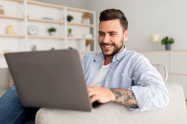 Modern job lifestyle. Happy tattooed man sitting on sofa at home and working on laptop — Stockfoto