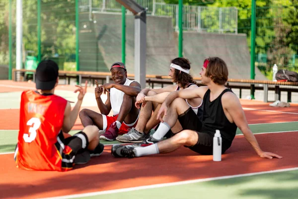 Multiracial basketball team resting and talking to each other on outdoor arena after game — Foto de Stock