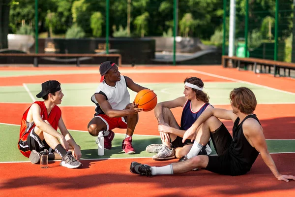 Team of young basketballers discussing their game strategy, sitting at outdoor stadium after training, free space — Foto de Stock