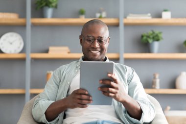 Happy black mature man using digital tablet, sitting in armchair over shelves with accessories, reading book online