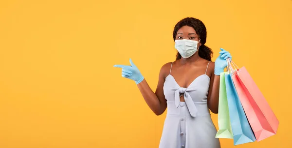 Young black lady holding gift bags, wearing face mask and rubber gloves, pointing at empty space on orange background — Stock fotografie
