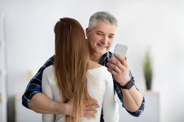 Mature man embracing his wife, stuck in smartphone, not paying attention, texting his lover, going through family crisis — Stock fotografie