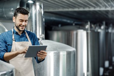 Young man work at beer factory. Brewer, industrial equipment and modern smart digital device at brewery