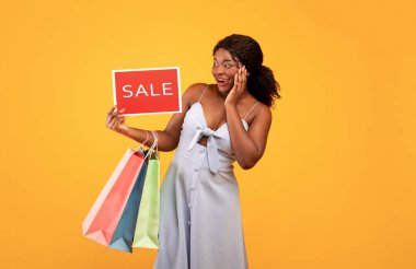 Pretty African American woman holding shopping bags and SALE sign, feeling excited over orange studio background