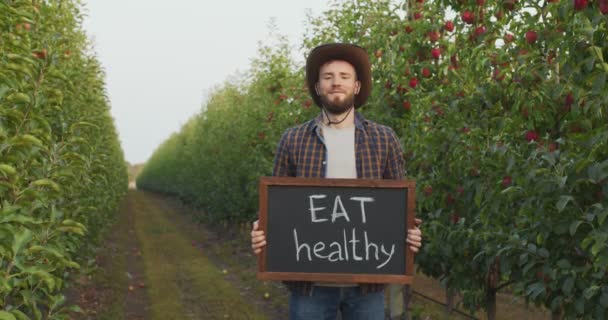 Młody rolnik pokazujący planszę Jedz zdrowo do kamery i uśmiechnij się, pozując na plantacji jabłek z dojrzałymi czerwonymi jabłkami na drzewach — Wideo stockowe