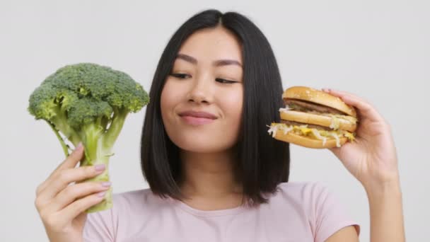 Китаянка держа брокколи и бургер выбирая питание, белый фон — стоковое видео