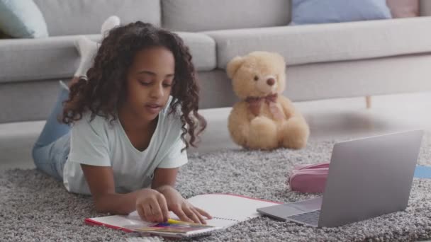 Мила афроамериканська дівчина малює з фетром, лежить на підлозі з ноутбуком, дивиться онлайн уроки мистецтва, повільний рух — стокове відео