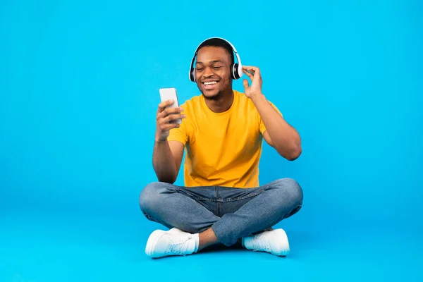 African Guy ouvir música usando Smartphone usando fones de ouvido, fundo azul — Fotografia de Stock