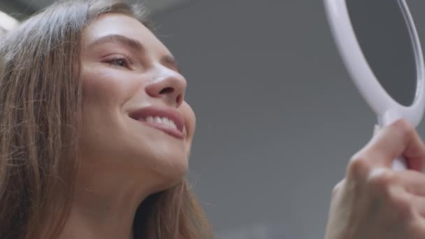 Mutlu genç kadın aynaya bakıyor, tedaviden sonra mükemmel gülümsemesinin tadını çıkarıyor ve dişçilikte beyazlatma prosedürlerini uyguluyor. — Stok video